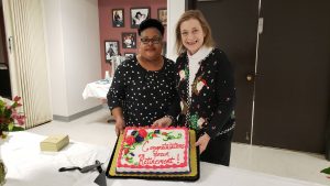 African American woman and Caucasian woman smiling behind a table while holding up a cake. Cake reads Congratulations On Your Retirement.