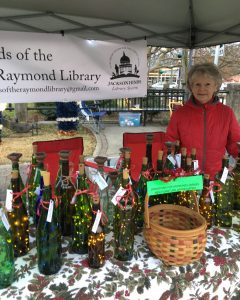 Caucasian woman with red coat standing behind table with decorative bottles. Sign Reads Friends of the Raymond Library.