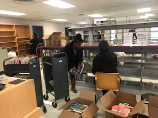 Caucasian man sitting down and African American woman standing while moving books from rolling cart to bookshelf.