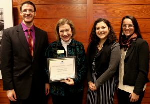 Group picture of one Caucasian man and three Caucasian women in business attire smiling. First Caucasioan woman holding up an award certificate.