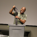 African American man with khaki T-shirt smiling and removing a black snake from a plastic gray container.