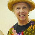 Headshot of older Caucasian woman smiling and wearing a straw hat, metal earrings and multicolored clothes