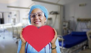 smiling Caucasian woman wearing surgical scrubs and holding large cutout of red heart. Hospital room background.