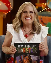Smiling Caucasian woman with blonde hair, glasses and while blouse holding a children's book