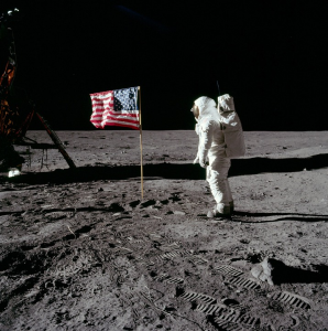 astronaut on moom to the right of the American flag. Spacecraft in background on left.