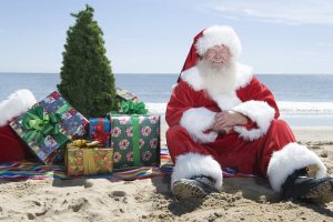 Happy senior Santa Claus sitting on beach with presents and Christmas tree