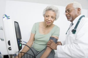 older African American male doctor and seated older African American woman wearing a blood pressure cuff. Both are looking at the screen of a blood pressure machine.